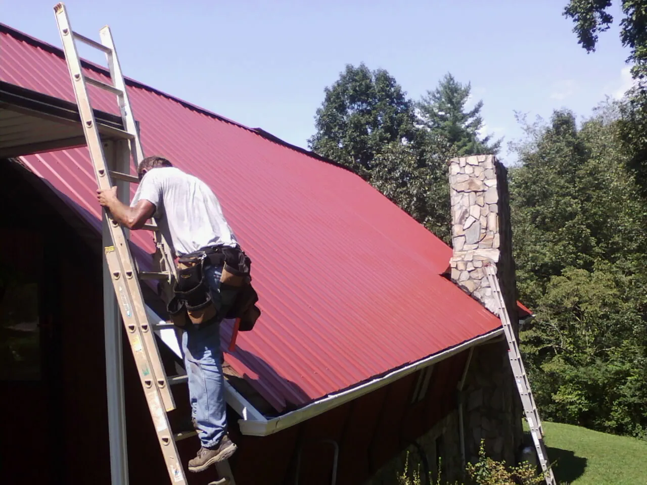 Roofer on a ladder beside a red metal roof and stone chimney — Soffit & Fascia Repair by Roofing Brothers in New Windsor